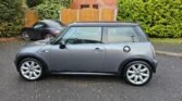 A gray Mini Cooper S is parked on a residential street. The car has silver alloy wheels and a black roof. In the background, there is another black car parked near a wooden fence, with trees and bushes lining the surroundings. The pavement is slightly wet, indicating recent rain.