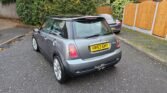 A silver Mini Cooper with the license plate HV53 GHY is parked on a paved street. This r53 model features a black roof and dual exhaust pipes. The area is surrounded by greenery and wooden fences, while fallen leaves scatter the ground. Another gray vehicle is partially visible in the background.