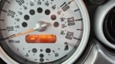 Close-up of a Mini Cooper's analog speedometer, showing a maximum of 150 mph or 240 km/h. The needle points to approximately 0 mph. Below, an orange digital odometer displays 086967 miles. Nearby are a hazard light button and an air vent. The speedometer and controls are set into a sleek, gray dashboard.