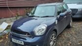 A dark blue Mini Cooper Countryman SD is parked on a gravel surface next to a large white vehicle. The car's license plate reads "SE65 KUN." The area is surrounded by leafless trees, a corrugated metal fence, and various pieces of debris on the ground. The sky appears overcast.