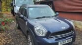 A dark blue Mini Countryman Cooper SD with the license plate SE65 KUN is parked on a gravel driveway next to a brown building. Behind it, a tow truck with an orange light on its roof is visible. The scene is set on a cloudy day with bare trees in the background, suggesting potential spare or repairs ahead.