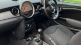 Interior of a compact MINI showcasing the dashboard and front seats. The dashboard features a large central speedometer and a steering wheel on the right side, indicating a right-hand drive. The interior is predominantly black with fabric seats and silver accents. A residential street is visible through the windows.