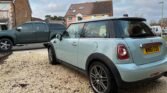 A light blue MINI Cooper with the registration plate "WN61 EOD" is parked on a gravel driveway in front of a two-story house. A dark grey vehicle is adjacent to it. The sky is mostly cloudy with some patches of blue and pink tones. The street and surrounding houses are visible in the background.