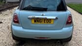 A light blue MINI Cooper is parked on a gravel driveway. The car's rear view shows a UK license plate reading "WN61 EOO", silver trim, and a rear window with some objects visible inside. Surrounding the car are suburban houses with brick facades and driveways.