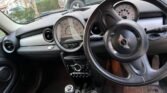 Interior view of a MINI Cooper vehicle showing the steering wheel with the distinctive MINI logo, a manual gear shift, and the dashboard. The dashboard features a large central speedometer and various control knobs below it. Outside the car, a green vehicle and some foliage are visible.