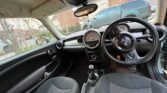 Interior of a MINI featuring a black steering wheel, a central dashboard with two large circular speedometers, and controls below. The car has black seats and door panels, with the driver's side door open. Houses, a tree, and a green vehicle are visible through the windows outside.