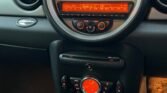 View of a MINI car dashboard featuring a large, circular speedometer with orange lighting. Below it, there is an illuminated radio display and control buttons, climate control dials, and various switches. The sleek interior is black, with a gear shift knob visible at the bottom of the image.