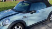 A light blue MINI Cooper convertible is parked on a residential street. The car features a black soft top roof and black racing stripes along the hood. In the background, there are rows of brick terraced houses with pitched roofs, small front gardens, and a well-maintained grassy area.