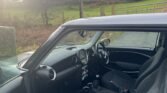 The image shows the interior of a Mini One car parked on a rural road. The photo, taken from the driver's side, captures the front seats, dashboard with graphite details, steering wheel, and gear shift. The window is slightly open, revealing a grassy field with trees and fencing in the background.