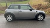 A grey Mini One compact car is parked on a damp road with a rural, grassy landscape in the background. The car has a sleek design with black trim around the wheels and windows, five-spoke alloy wheels, and runs on petrol. The overcast weather adds a muted graphite tone to the scene.