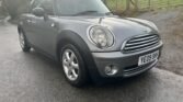 A gray MINI Cooper is parked on a wet road. The car has silver alloy wheels and a chrome-bordered grille. The front license plate reads "YE09 OHH." In the background, there is a lush green field separated by a wire fence, and distant trees are visible under a cloudy sky.