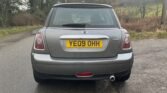 A silver Mini One car is parked on a rural road, shown from the rear. The UK license plate reads "YE09 OHH." The petrol-driven car features distinctive oval taillights, a rear window wiper, and a single exhaust pipe on the right side. Trees and grasslands are visible in the background.