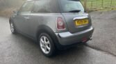 A graphite MINI One with a yellow rear license plate reading "YE09 OHH" is parked on a wet road. The car has two doors and a black roof. In the background, there is a metal gate and a grassy field with trees and bushes. The sky appears cloudy.