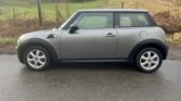 A silver Mini One is parked on a wet road beside a grassy field with a fence and trees in the background. The car's side profile shows its compact size, with the driver's side door, four wheels, black trim, and black side mirrors all visible. The sky is cloudy, suggesting recent or impending rain.