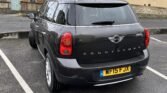 A black MINI Countryman with license plate "WF15 FJX" is parked in an empty parking lot. The vehicle is positioned next to a large stone building with tall windows. The ground appears to be wet, suggesting recent rain. The rear view of the car showcases its brand logo and model name.