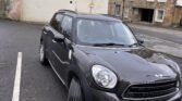 A black MINI Cooper is parked in a nearly empty parking lot next to a curb. It is positioned at an angle, facing slightly to the left. The background includes a few multi-story buildings with brick and stone facades, windows, and some graffiti on the walls. The car has a visible UK license plate.