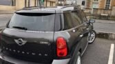 A black MINI Countryman Cooper D is parked at an angle in a near-empty parking lot with various beige and stone buildings in the background under an overcast sky. The vehicle's license plate reads WF15 FJX. The street is quiet, and traffic lights are visible.