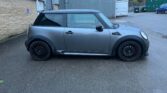Side view of a grey MINI Cooper S hatchback with a black roof and black wheels, parked on a wet asphalt surface near a corrugated metal building and green storage containers. The background includes a grassy hill and overcast sky, emphasizing the well-used look of the MINI.