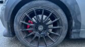 Close-up of a MINI's front wheel, displaying a black multi-spoke alloy rim with a red brake caliper behind it. The tire is mounted on a grey car, with visible dirt and grime on the wheel and the lower part of the car body. The wheel rests on a rough asphalt surface.