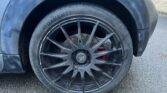A close-up view of a MINI's right front black alloy wheel with a low-profile tire. The wheel features multiple thin spokes radiating from the central hub, with a visible disc brake and red brake caliper behind it. The car's black fender and asphalt road are also visible.