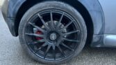 Close-up of a MINI's black alloy wheel with thin spokes, featuring a visible red brake caliper behind it. The tire is mounted on a gray-colored vehicle, with some dirt and wear marks visible on the wheel arch and body. The backdrop is a rough-textured asphalt surface.