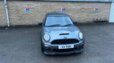 A gray MINI Cooper is parked in front of a tan brick wall with a corrugated metal roof. The car's license plate reads "Y2 THH." Two signs on the wall indicate designated wheelchair-accessible parking spots. The ground is paved and slightly wet, suggesting recent rain.