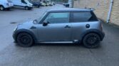 A gray MINI Cooper with black rims is parked in a lot near industrial buildings. The vehicle is photographed from the side, showcasing its compact size and sporty appearance. The sky is overcast, and the ground appears slightly wet. Other vehicles and buildings are visible in the background.