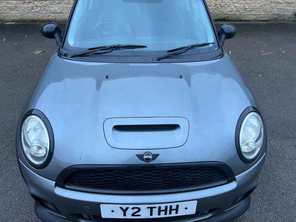 A gray MINI Cooper is parked on an asphalt surface. The front of the car is visible, including its headlights, hood, grille, and side mirrors. The license plate reads "Y2 THH." The background features a stone wall.
