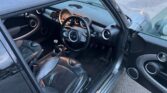 The image shows the interior of a compact MINI with black leather seats and a modern dashboard. The steering wheel features a central emblem, and the car has manual transmission. The driver's side door is open, revealing part of the parking lot outside. The interior is sleek and minimalistic.