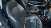 The image shows the front interior of a mini car with two black leather seats, both showing some wear and creasing. The right-hand drive layout, visible dashboard controls, and rounded door design with a silver handle are classic mini features.