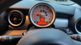 The image shows the dashboard of a MINI car, featuring a central circular speedometer with red backlighting, displaying a speed of 0 mph, and an odometer reading of 78089 miles. The surrounding dashboard includes vents and control stalks. The windshield appears to have raindrops on it.