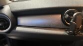 The image shows the interior of a MINI car, specifically the dashboard area. The dashboard has a sleek, black design with a carbon-fiber textured strip in the center. Air vents are present on both ends, and a phone holder is attached to the right side air vent. Raindrops are visible on the windshield.