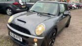 A grey MINI Cooper with registration plate "GL06 FFD" is parked in a lot. The car features a black grille, black side mirrors, and black wheels. Its headlights are on, and several other cars and buildings are visible in the background. The ground is covered in gravel.