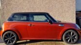 A bright orange MINI Cooper is parked on a concrete surface in front of a textured beige wall. The car boasts a black roof and black alloy wheels. Sunlight casts a shadow on the lower part of the wall and highlights the glossy finish of this stylish vehicle.