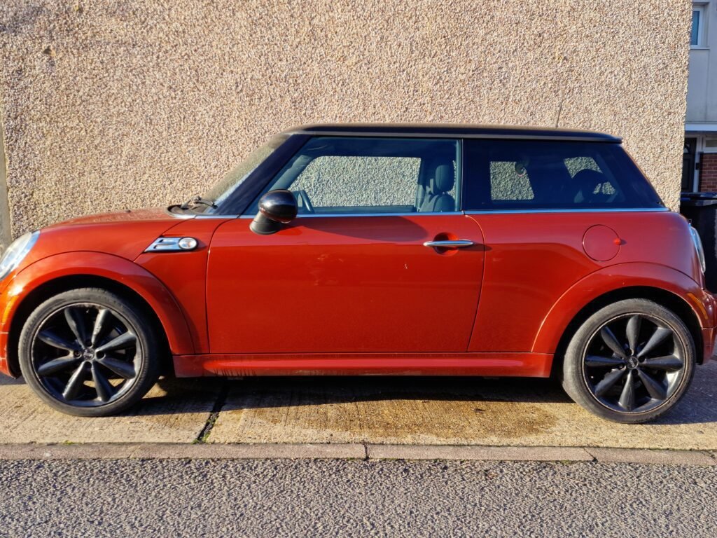 A red MINI Cooper with black alloy wheels is parked on a concrete driveway next to a beige textured wall. The car's design includes a black roof and side mirrors, characteristic of the MINI Cooper style. The lighting suggests that it is a sunny day, as indicated by the shadows.