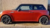 A red MINI Cooper with black alloy wheels is parked on a concrete driveway next to a beige textured wall. The car's design includes a black roof and side mirrors, characteristic of the MINI Cooper style. The lighting suggests that it is a sunny day, as indicated by the shadows.