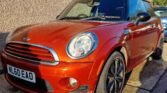 A bright orange MINI Cooper with a UK license plate reading "NL60 EAO" is parked by the side of a textured stone building. The car boasts black alloy wheels, a black roof, and chrome accents. The sun casts shadows, highlighting its glossy finish. In the background are another building and part of another vehicle.