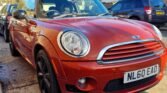 A red MINI Cooper with a black roof is parked on a residential street. The car's front license plate reads "NL60 EAO." Other vehicles and houses are visible in the background. The sun is shining, casting shadows on the ground. The MINI has a sleek design with distinctive front headlights.