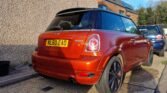 A shiny, orange MINI Cooper with black wheels is parked on the right side of a driveway next to a pebble-dash wall. The vehicle's license plate reads "NL60 EAO." In the background, another car and a modern brick house with solar panels on the roof are visible. The sky is clear and blue.