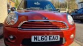 A red MINI Cooper is parked on a residential street. The front view of the car showcases its trademark rounded headlights, chrome grille, and a license plate reading "NL60 EAO." Surrounding the MINI are suburban houses, with leafless trees in the background under a partly cloudy sky.