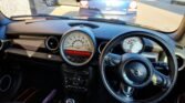 Interior of a MINI Cooper, showcasing the steering wheel with the iconic MINI logo, a large central speedometer with digital and analog displays, and a tachometer to the right. The dashboard is sleek and minimalist, with two vehicles visible through the windshield.