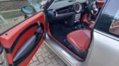 The interior of a beige MINI Cooper is shown with the driver's side door open. The interior features a combination of red and black upholstery with red leather seats and door panels. The dashboard, steering wheel, and center console are also visible. The car is parked on a brick driveway.