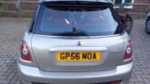 A rear view of a silver MINI Cooper with the license plate "GP56 MOA." The car is parked on a brick driveway next to a brick building. The MINI's rear window and taillights are visible, along with twin exhaust pipes at the bottom. The driver's side door is slightly open.