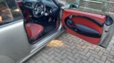 A MINI with its driver's side door open reveals a black and red interior. The seat is red with black accents, and the door panel is red with a black handle. The steering wheel and dashboard are visible, and the car is parked on brick pavement in front of a brick wall with windows.