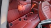 Interior view of a MINI with red leather car seat setup. The front passenger seat is fully visible, while the backseat shows two headrests and is designed to seat two passengers. The seats feature horizontal stitching and a central armrest with cup holders. The side door panel is partially visible.