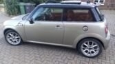 A silver MINI Cooper with a black roof is parked on a brick driveway. The car has a sunroof, alloy wheels, and a fuel cap on the rear side panel. Behind the car, there is a garden with green shrubs partially blocked by gray and green trash bins. The exterior is clean and well-maintained.