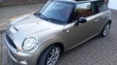 A metallic beige MINI Cooper S is parked on a brick driveway. The car features a black roof with a sunroof, chrome side mirrors, and sporty alloy wheels. In the background, there's a brick building with white-framed windows and a partially open garage door.