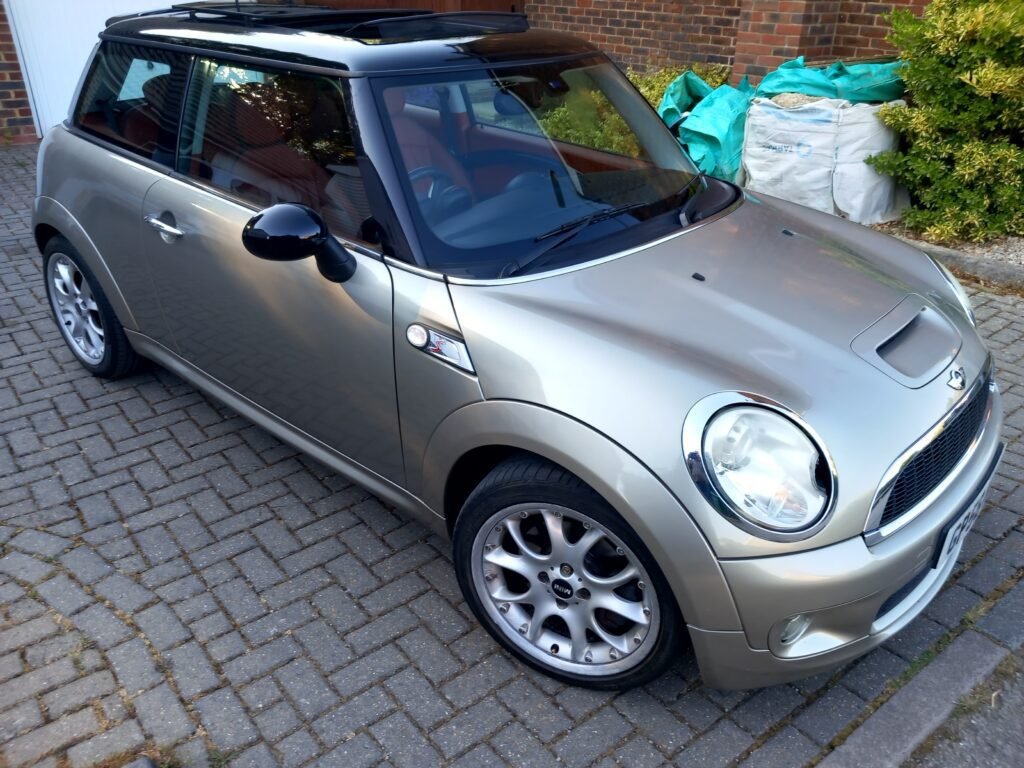 A silver MINI Cooper hatchback is parked on a brick driveway. The car features a black roof with a sunroof, shiny alloy wheels, and side mirrors with a black finish. In the background, there is a brick wall, some green shrubbery, and several covered items. The car appears clean and well-maintained.