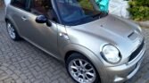 A silver MINI Cooper hatchback is parked on a brick driveway. The car features a black roof with a sunroof, shiny alloy wheels, and side mirrors with a black finish. In the background, there is a brick wall, some green shrubbery, and several covered items. The car appears clean and well-maintained.