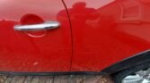 A red MINI with a noticeable scratch on its side door. The car door features a chrome handle. The bottom of the car is lined with black trim. The reflection of a person taking the photo and nearby structures can be seen on the shiny surface. The vehicle is parked on a brick and gravel surface.